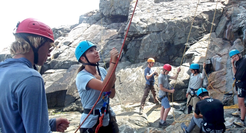 A group of people wearing safety gear stand at the base of a rock wall and belay climbers. 